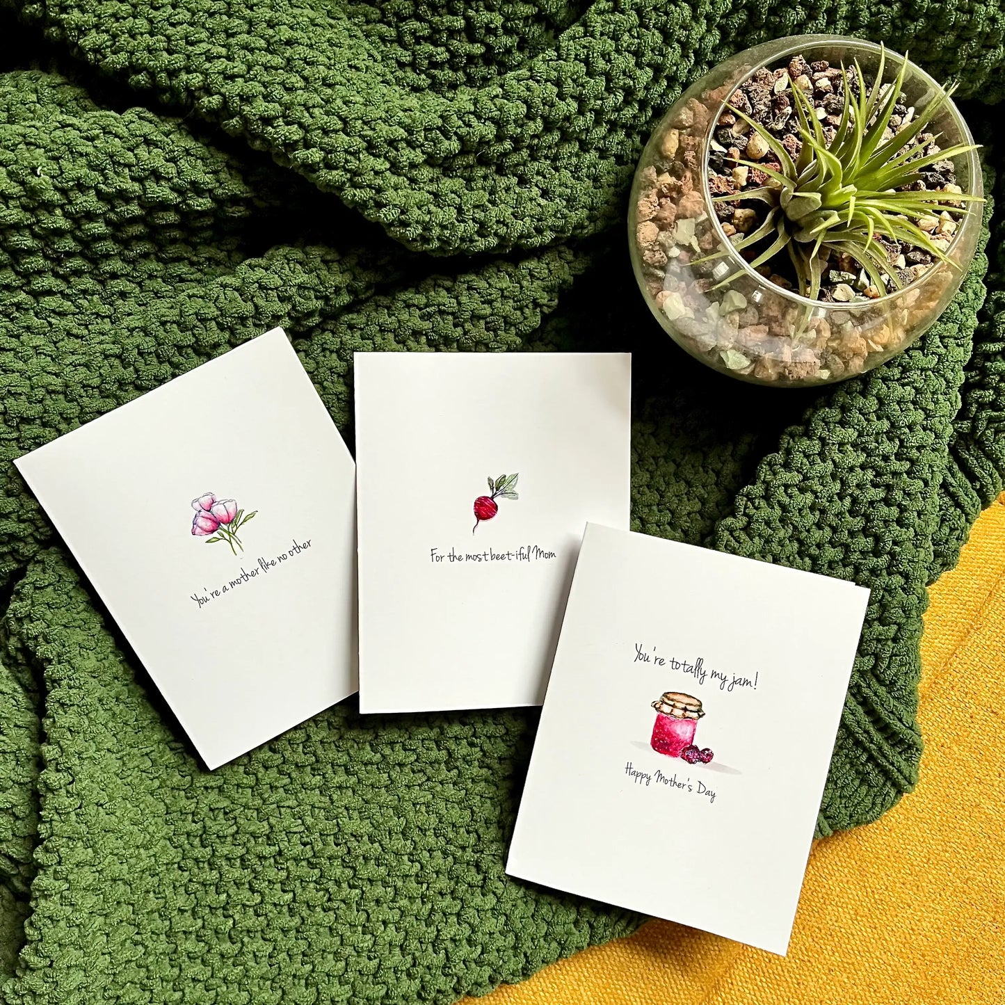 Handcrafted Mother's Day cards with watercolor illustrations on green knitted fabric next to a succulent plant.