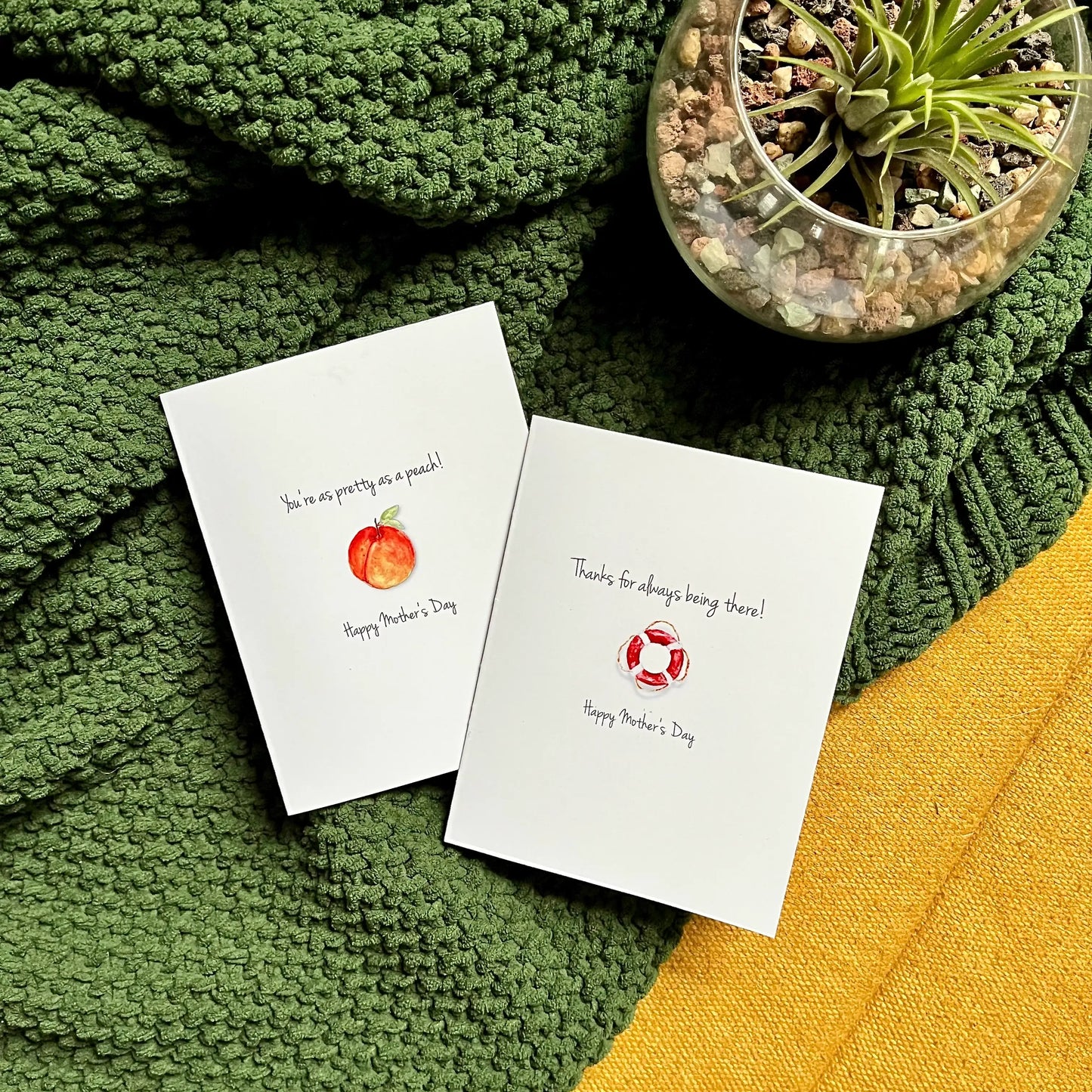 Two handcrafted Mother's Day cards with original artwork on a knitted green blanket beside a potted succulent.