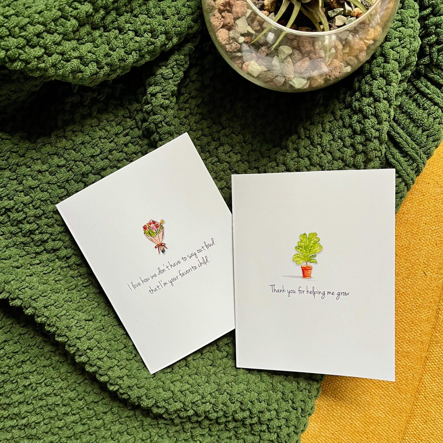 Two handcrafted greeting cards with floral and plant illustrations on a textured green blanket beside a glass bowl with succulents.
