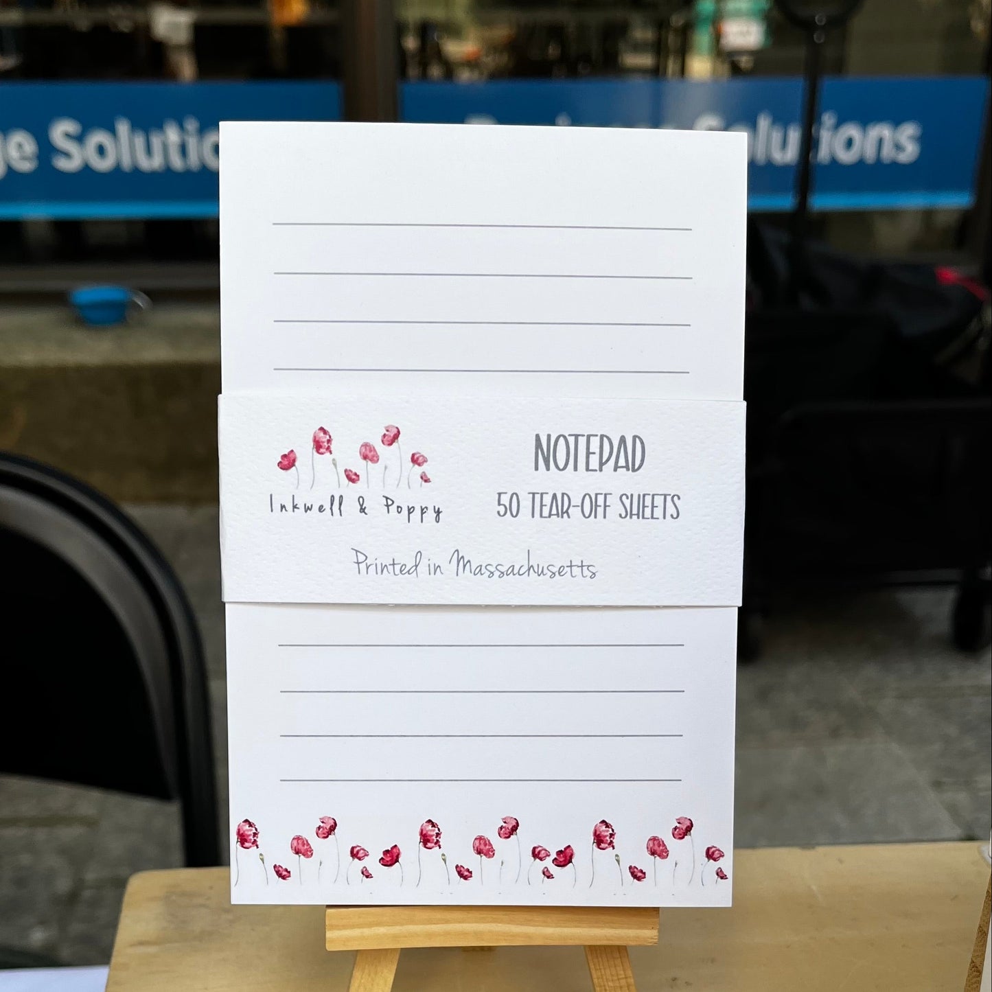 A white notepad displayed on a small wooden easel, with printed text reading 'Notepad 50 Tear-Off Sheets' and a small red floral border. The setting appears to be a real indoor environment with furniture and a window in the background.