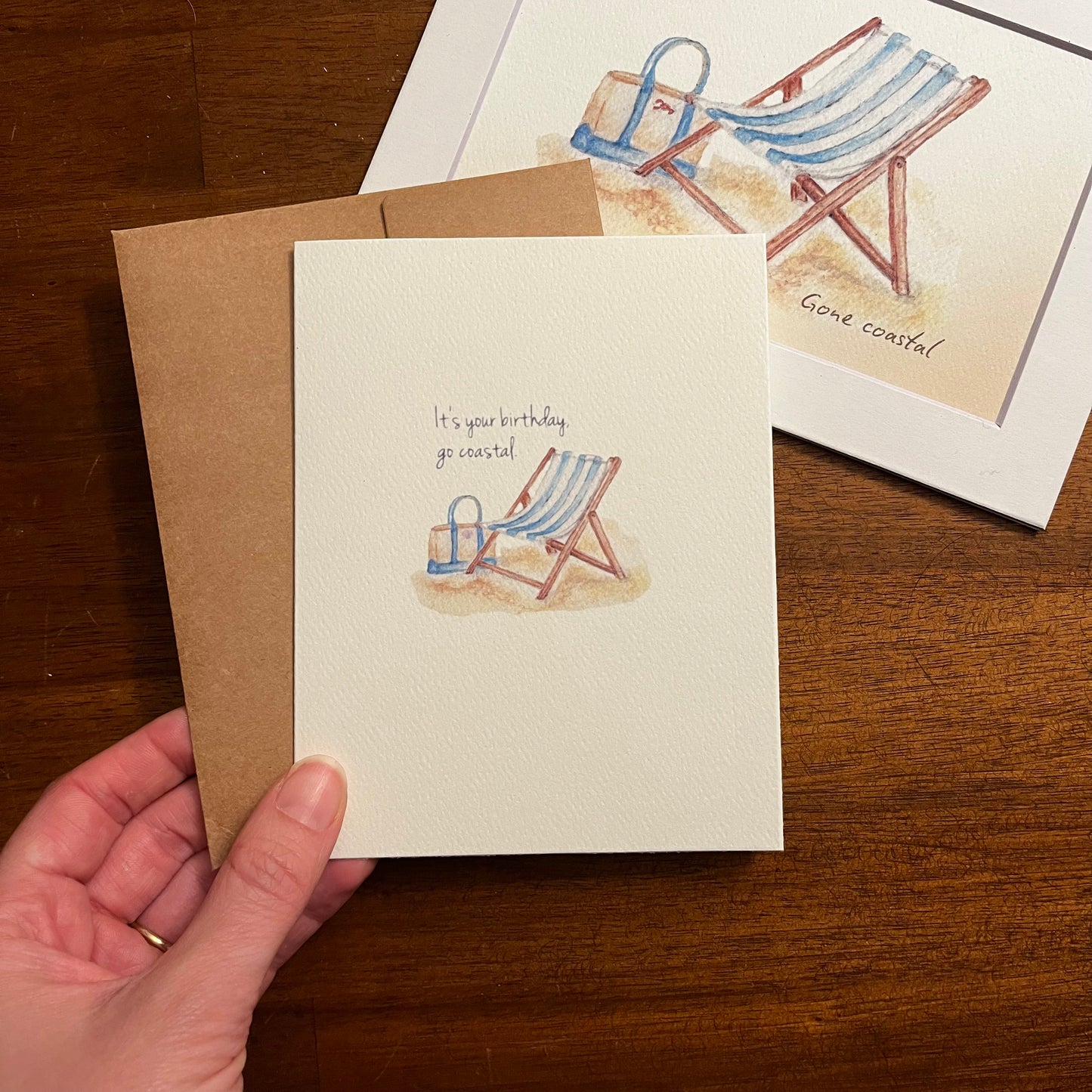 Hand holding a coastal-themed greeting card with a beach chair and bucket illustration on a wooden surface.